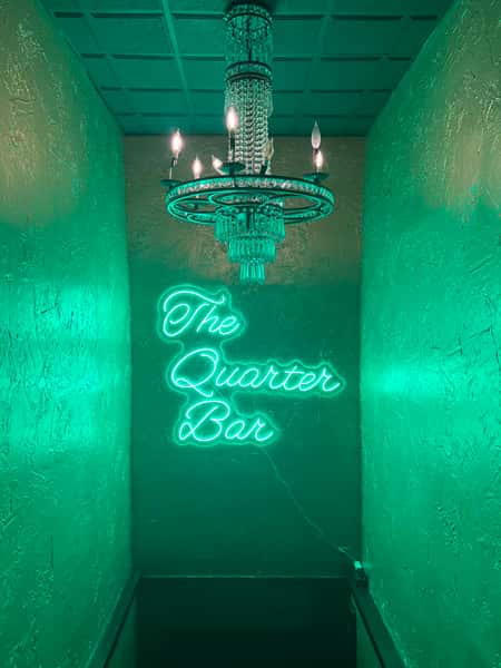 Chandelier-lit hallway with green neon sign reading "The Quarter Bar" against textured walls.