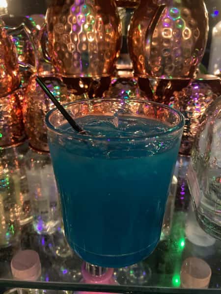 Bright blue cocktail served over ice in a clear glass with a black straw, surrounded by copper mugs and colorful reflections.
