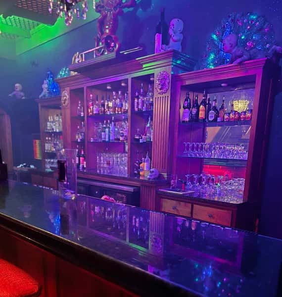 Eclectic bar setup with colorful lighting, vintage wooden shelves stocked with liquor, and quirky doll decorations throughout.