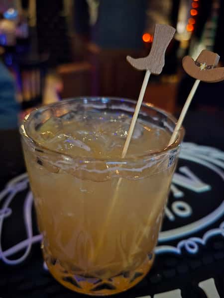 A cocktail on the rocks in a textured glass, garnished with skewers topped with wooden cowboy boot and hat decorations.