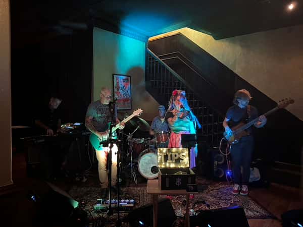 Live band performing on stage in a dimly lit venue with a lit "TIPS" box at the front and staircase in the background.