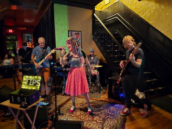Live band performing indoors with a female vocalist, guitarists, and drummer, next to a glowing "TIPS" sign and tip box.