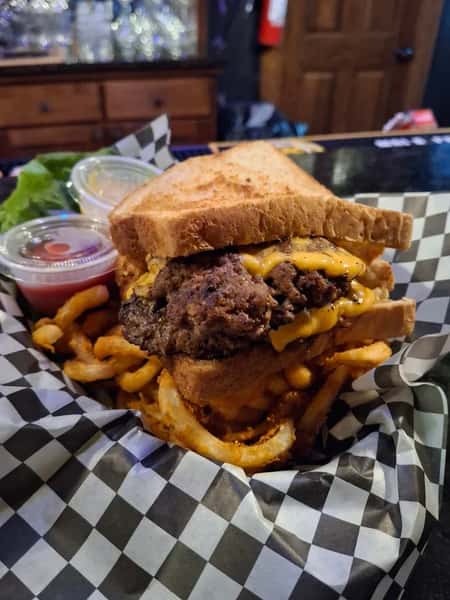 Cheeseburger on toasted sandwich bread with curly fries, served in a checkered basket with ketchup and sauce cups on the side.