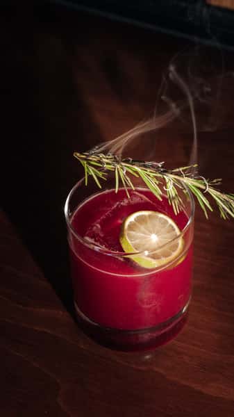Red citrus cocktail garnished with rosemary and a lemon slice on a wooden table, a stylish Italian restaurant drink.