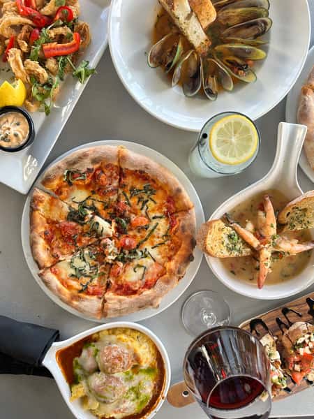 Overhead view of an Italian restaurant table featuring a wood-fired pizza with melted mozzarella and roasted cherry tomatoes, steamed mussels in broth with toasted bread, crispy calamari with peppers, shrimp scampi with garlic bread, baked meatballs in marinara and pesto, and bruschetta, served with red wine and lemon water for a Mediterranean seafood dining spread.