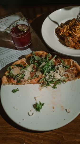 Prosciutto and arugula pizza with shaved parmesan on a plate beside spaghetti and meatballs and a rosemary-garnished cocktail at a cozy Italian restaurant table.