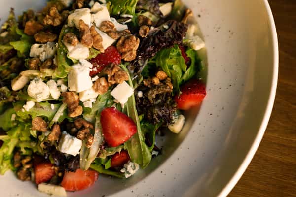 Fresh mixed greens salad with strawberries, candied walnuts, blue cheese crumbles, and sliced almonds in a large white bowl.