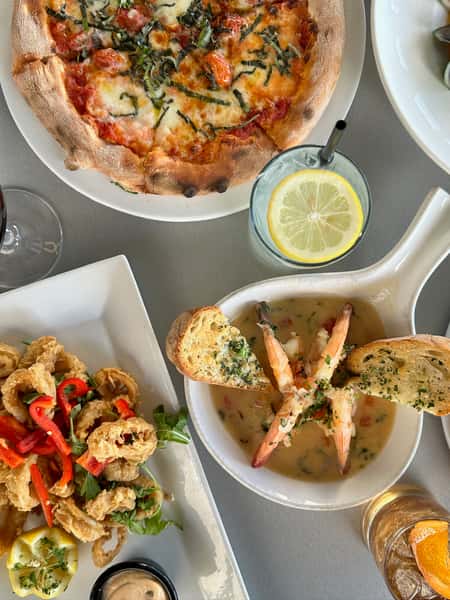 Overhead table spread featuring crispy calamari with peppers, shrimp scampi with garlic bread, margherita-style pizza, and lemon water.