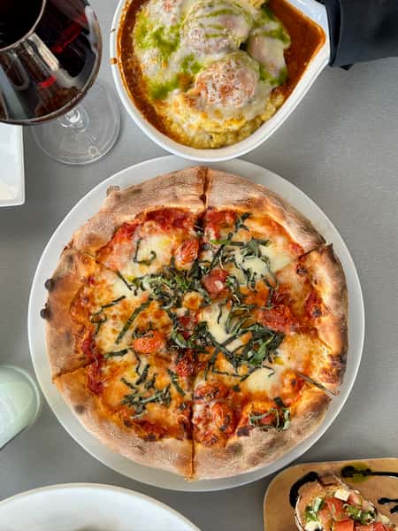 Wood-fired pizza with melted mozzarella and basil served alongside baked Italian meatballs with marinara and pesto and a glass of red wine.