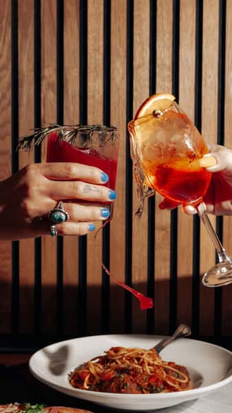 Two hands clinking bright Italian cocktails—an Aperol spritz and a red citrus cocktail—above a plate of spaghetti and meatballs.