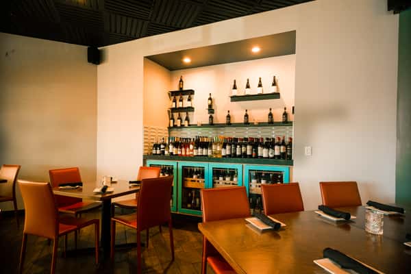 Modern Italian restaurant dining room with wooden tables, leather chairs, and a wine display bar lined with bottles, creating a warm upscale ambiance.