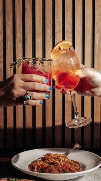 Two hands clinking vibrant Italian cocktails—an Aperol spritz and a red citrus cocktail garnished with rosemary—above a plate of spaghetti and meatballs at a stylish Italian restaurant table.
