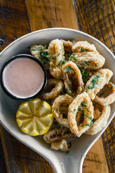 Crispy fried calamari served with lemon wedge and creamy dipping sauce in a white bowl on a rustic wooden table, a popular Italian seafood appetizer.