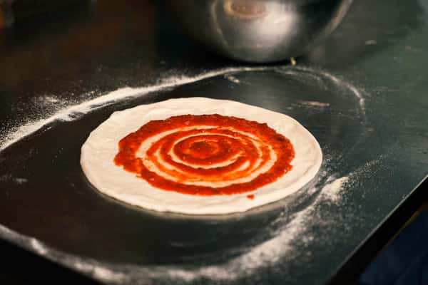 Fresh pizza dough topped with spiral tomato sauce on a floured prep station, capturing the start of a classic Italian wood-fired pizza.