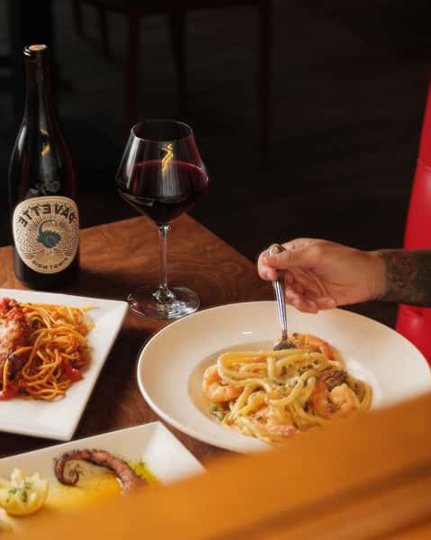 Italian dinner table with shrimp pasta in cream sauce, spaghetti marinara, grilled octopus, and a glass of red wine beside a wine bottle.