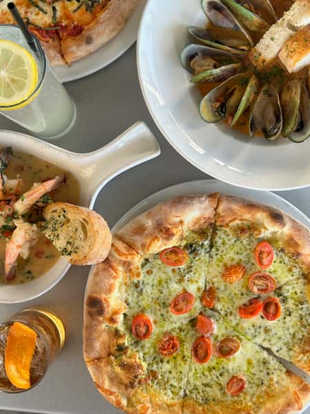 Overhead view of an Italian restaurant table with wood-fired pesto pizza, steamed mussels with toasted bread, shrimp scampi, and cocktails.