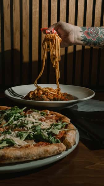 Hand lifting spaghetti with marinara and meatballs from a bowl beside a prosciutto arugula pizza on a restaurant table.