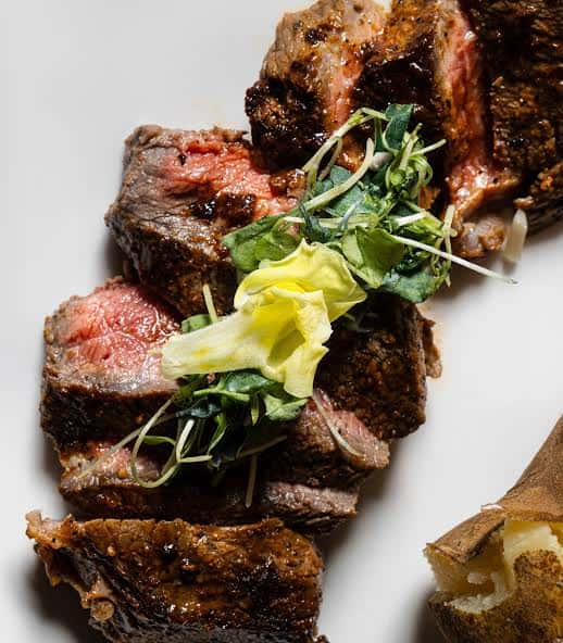 Sliced medium-rare steak with a seared crust, topped with microgreens and a yellow edible flower, served on a white plate alongside a piece of baked potato.