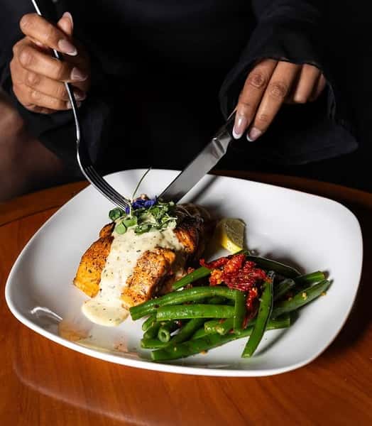 Blackened salmon fillet topped with creamy white sauce and microgreens, served with sautéed green beans and sun-dried tomatoes, on a square white plate as a diner cuts into the dish.