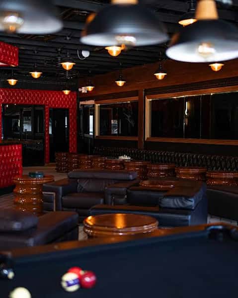 Stylish cigar lounge featuring plush leather seating, red tufted wall panels, black pool table with billiard balls, and warm pendant lighting for a refined social atmosphere.
