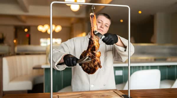 Chef presenting a hanging tomahawk steak in a restaurant dining area.