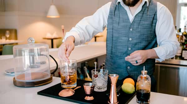 Bartender preparing cocktails behind a bar with bottles and citrus on the counter.