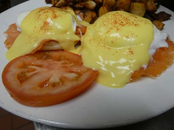 Smoked salmon eggs Benedict with poached eggs and hollandaise on an English muffin, served with tomato slices and breakfast potatoes.