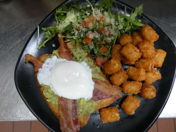 Brunch plate with avocado toast topped with bacon and a poached egg, crispy tater tots, and arugula salad with diced tomatoes.