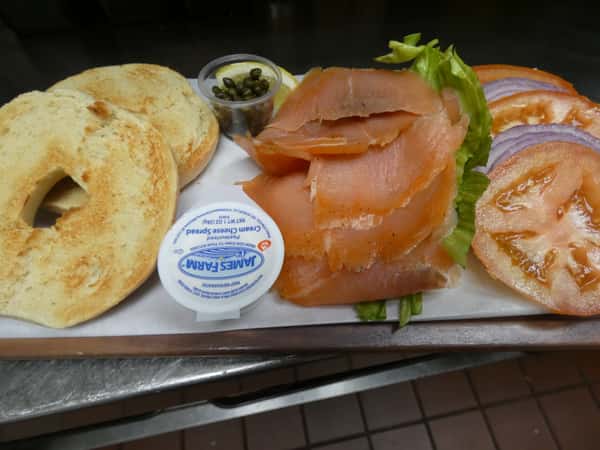 Toasted bagel with smoked salmon, cream cheese, capers, lettuce, tomato, and red onion, served deconstructed on a tray.