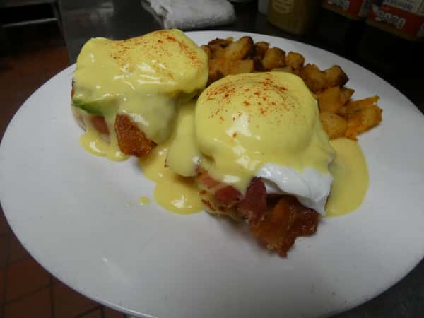 Classic eggs Benedict with poached eggs, bacon, avocado, and hollandaise sauce, served with a side of seasoned breakfast potatoes.