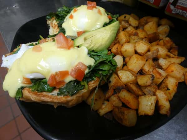 Vegetarian eggs Benedict with poached eggs, sautéed spinach, avocado, and hollandaise on toast, served with seasoned breakfast potatoes.