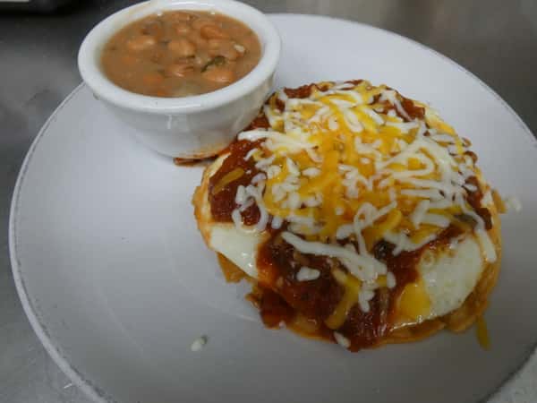 Huevos rancheros topped with red salsa and shredded cheese, served with a side of seasoned pinto beans in a small white ramekin.