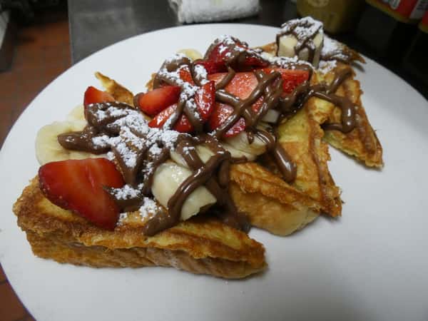 Thick-cut French toast topped with sliced strawberries, bananas, powdered sugar, and a generous drizzle of chocolate hazelnut spread.