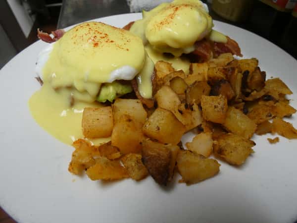 Eggs Benedict topped with creamy hollandaise and paprika, served over bacon and avocado with a side of golden, seasoned home fries.