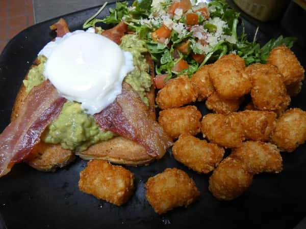Brunch plate with avocado toast topped with bacon and a poached egg, crispy tater tots, and arugula salad with diced tomatoes.