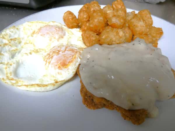 Country breakfast plate with two fried eggs, crispy tater tots, and chicken fried steak topped with creamy peppered country gravy.