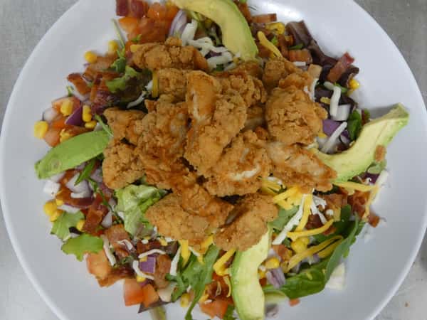 Fresh salad topped with crispy fried chicken, avocado slices, shredded cheese, bacon, corn, red onion, and chopped tomatoes.