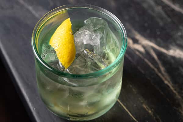 Clear spirit-forward cocktail in a short glass over large ice cubes, garnished with a lemon twist, served on a dark stone bar at La H