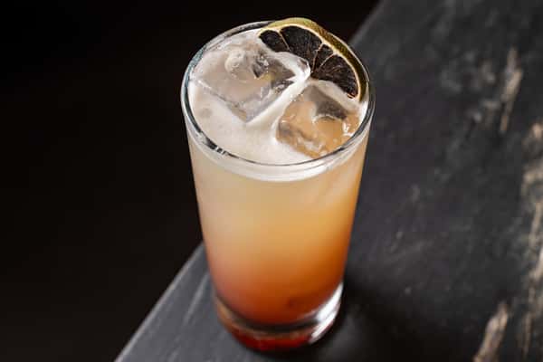 Refreshing tall mezcal cocktail with a gradient orange hue, served over ice and topped with a charred citrus wheel on a dark stone b