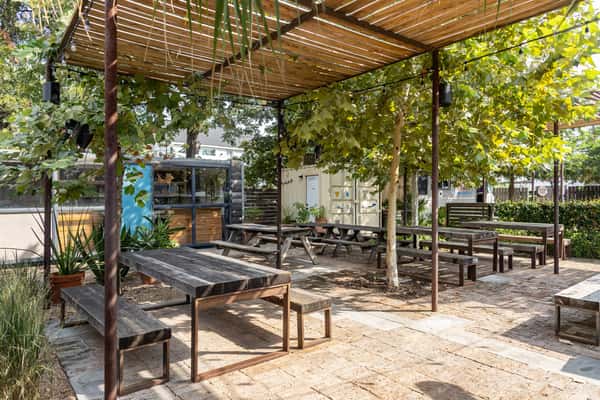 Outdoor courtyard at La Holly featuring wooden picnic tables under leafy trees and pergolas, with a view of nearby food trucks in a relaxed Austin setting.