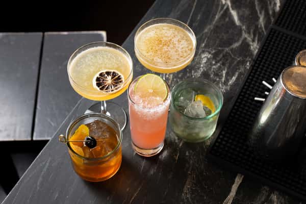 Assortment of five craft cocktails on a dark stone bar at La Holly, including mezcal and tequila-based drinks in coupe, highball, and lowball glasses with citrus and salt rim garnishes.