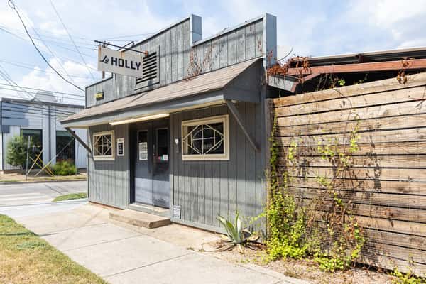 Corner view of La Holly cantina in East Austin, showcasing its rustic gray wood facade, vintage-style signage, and vine-covered fencing along a quiet street.