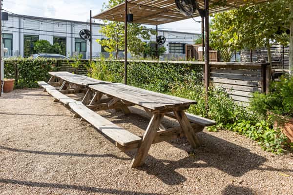 Outdoor patio at La Holly featuring rustic wooden picnic tables on gravel ground, shaded by string lights and greenery in a casual East Austin setting.