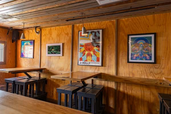 Cozy bar interior with wooden walls and ceiling, featuring vibrant Latin-inspired artwork and modern pendant lighting above high-top tables with black stools.