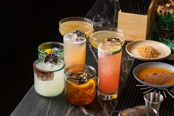 Vibrant lineup of six signature mezcal and tequila cocktails with citrus garnishes and salt rims, set on a dark marble bar with tools and ingredients nearby.