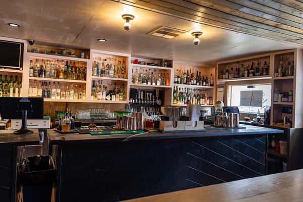 La Holly's bar interior featuring an expansive backbar of spirits and mezcal, black herringbone counter, and warm wood ceiling with modern lightin