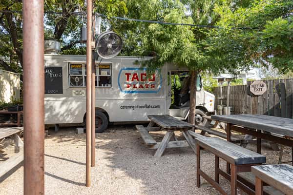 Taco Flats food truck parked in a shaded outdoor courtyard with rustic wooden picnic tables and gravel ground, offering casual al fresco dining under leafy trees.