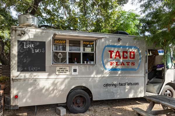 Close-up of the Taco Flats food truck parked under trees, with labeled order and pickup windows and bold red and blue branding, set in an outdoor dining courtyard