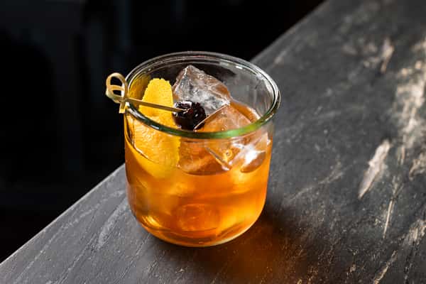 Classic Old Fashioned cocktail with orange peel and cherry garnish in a short glass, served over crystal-clear ice on a dark stone countertop.
