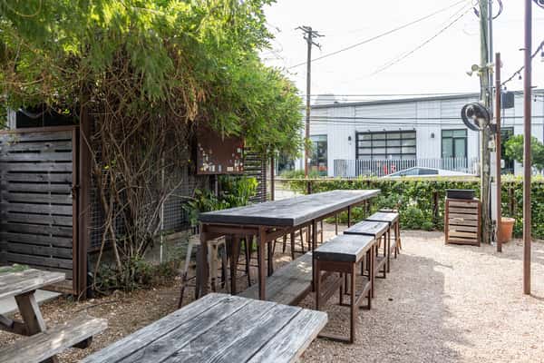 Outdoor courtyard at La Holly featuring high-top and picnic-style wooden tables on gravel ground, shaded by lush greenery and surrounded by an urban backdrop.